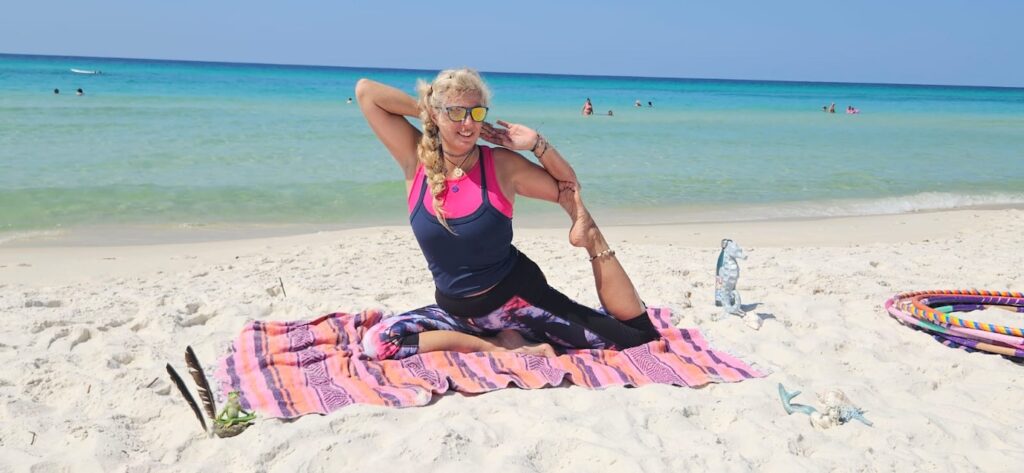 Eine Frau praktiziert Yoga in einer sitzenden Dehnposition auf einer bunten Decke am weißen Sandstrand von Panama City Beach, Florida, vor dem türkisfarbenen Meer.