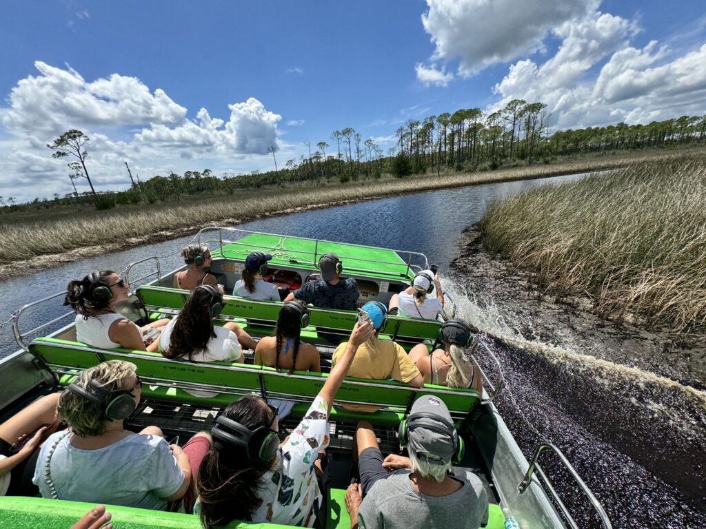 Eine Gruppe von Menschen sitzt auf einem grünen Airboat, das durch das Marschland von Panama City Beach, Florida, fährt, umgeben von Schilf und unter blauem Himmel.