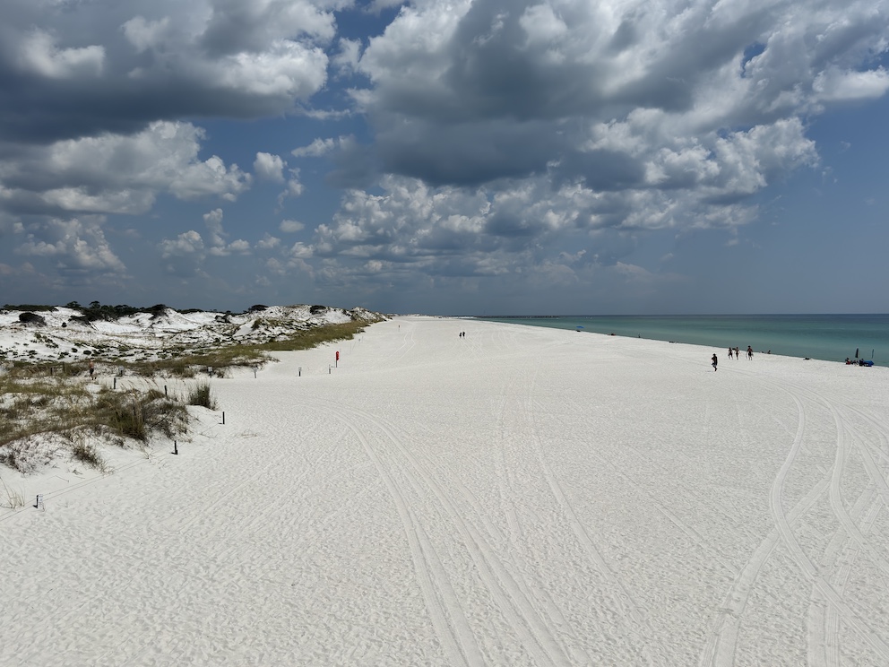 Ein weitläufiger, weißer Sandstrand in Panama City Beach, Florida, umgeben von Dünen und türkisfarbenem Wasser unter einer bewölkten, aber sonnigen Himmel.