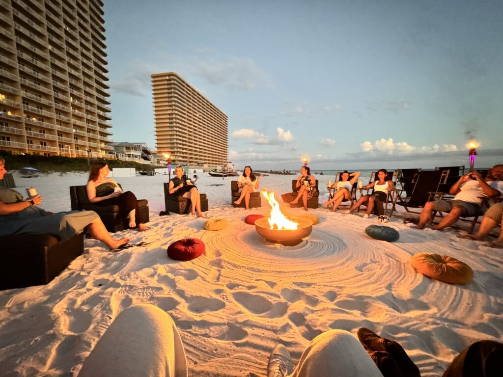 Menschen sitzen gemütlich um ein Lagerfeuer am weißen Sandstrand von Panama City Beach, Florida, bei Sonnenuntergang, umgeben von modernen Hochhäusern.