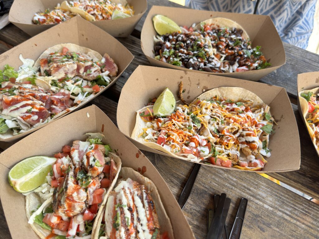 Verschiedene Portionen frischer Tacos, belegt mit Fisch, Gemüse und Sauce, serviert in umweltfreundlichen Pappbehältern auf einem rustikalen Holztisch.