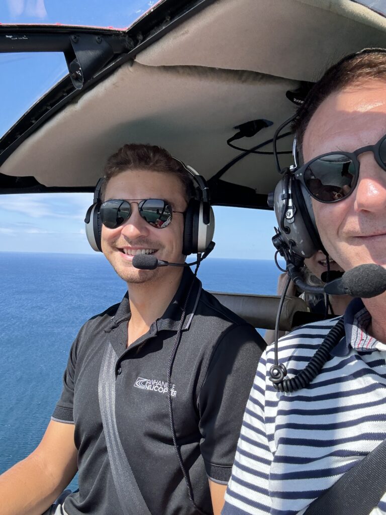 Ein Helikopterpilot und ein Passagier lächeln während eines Fluges über die Küste von Panama City Beach, Florida. Im Hintergrund ist das türkisfarbene Meer zu sehen.