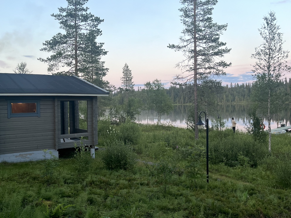 Holzsauna am Ufer eines Sees in Finnland, umgeben von Bäumen und grüner Natur in der Abenddämmerung.