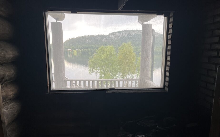 Blick aus dem Fenster einer Rauchsauna auf einen ruhigen See und bewaldete Berge in Finnland.