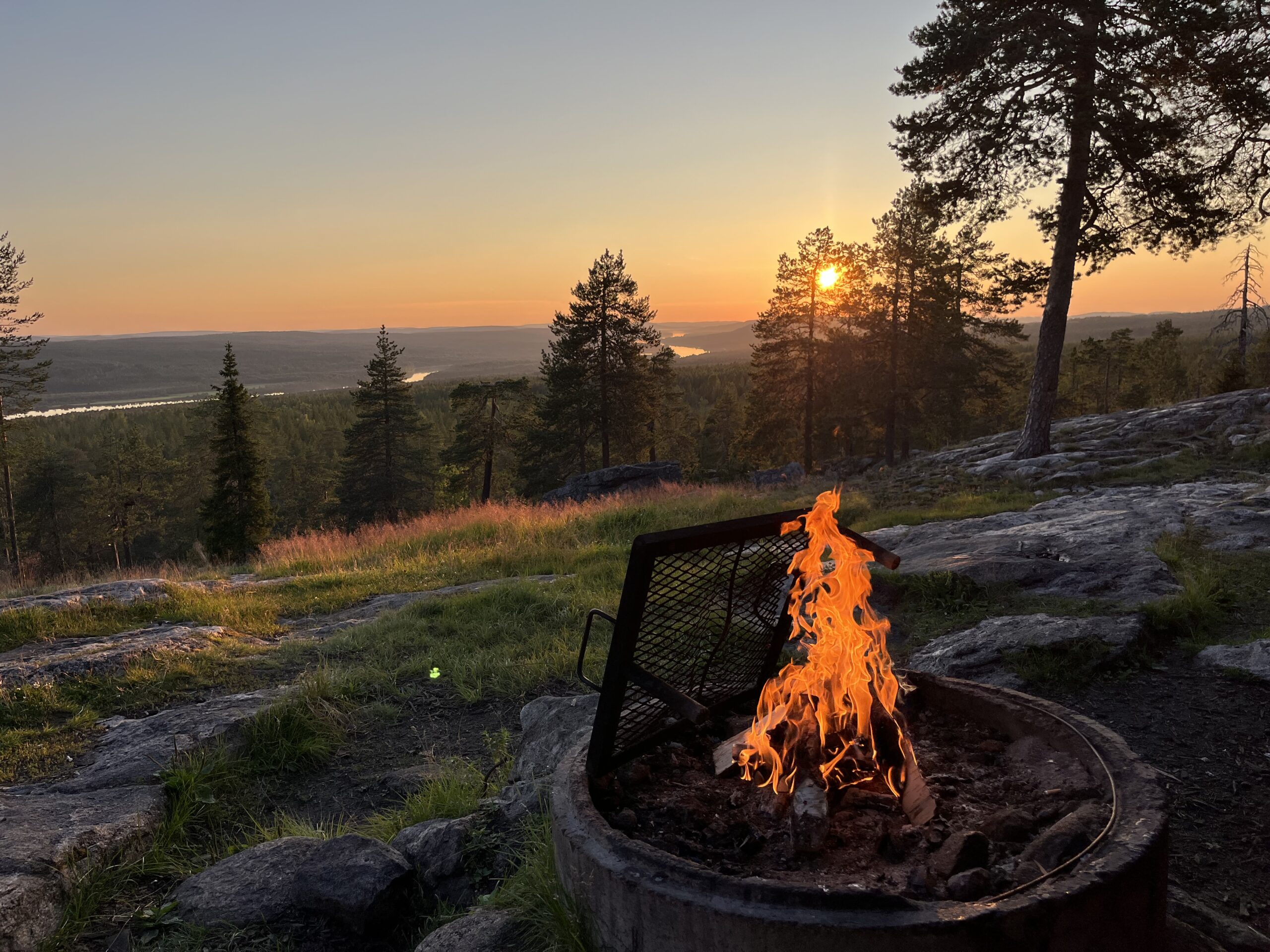 Lagerfeuer im Vordergrund, dahinter ein Sonnenuntergang über den bewaldeten Hügeln und Flüssen Finnlands.
