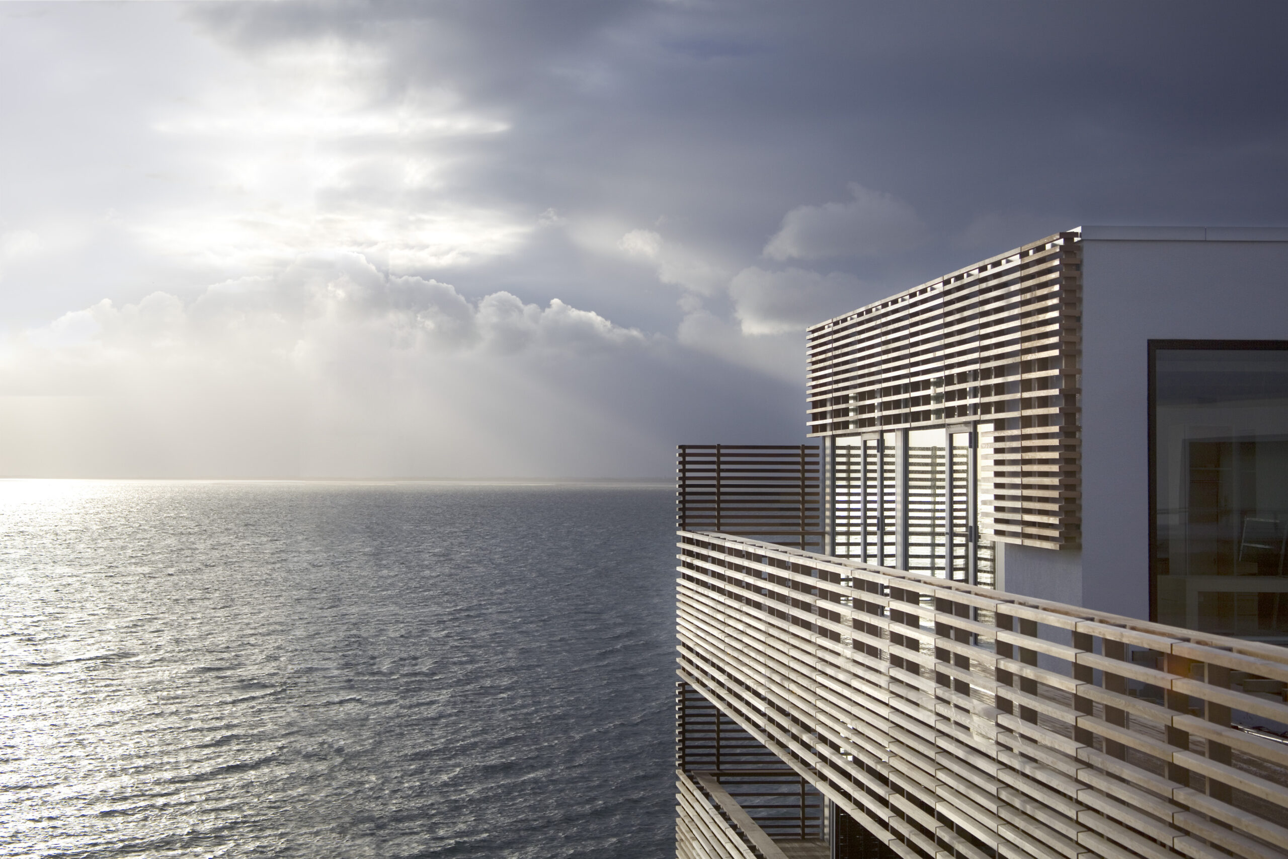 Modernes Gebäude mit Holzverkleidung auf Sylt, das direkt am Meer liegt, mit Aussicht auf die Nordsee und einen bewölkten Himmel, durch den Lichtstrahlen fallen.