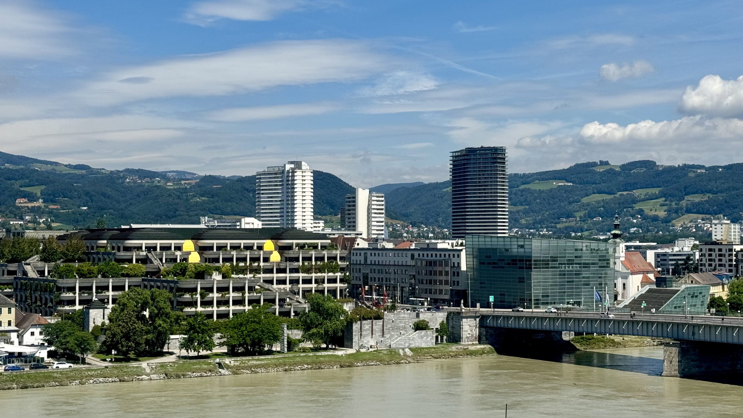 Skyline von Linz mit modernen Hochhäusern und Glasgebäuden, im Vordergrund die Donau und im Hintergrund bewaldete Berge unter blauem Himmel.