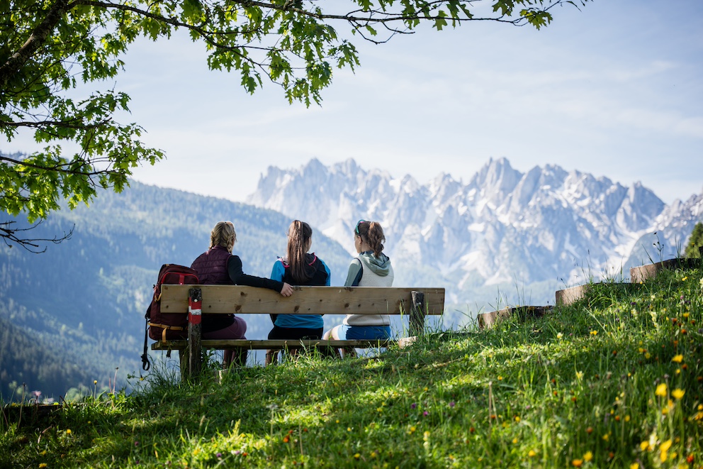 Auf diesem Bild sieht man drei Damen, die eine Rast auf einer Bank während einer Wanderung machen mit dem Blick auf den Gosaukamm