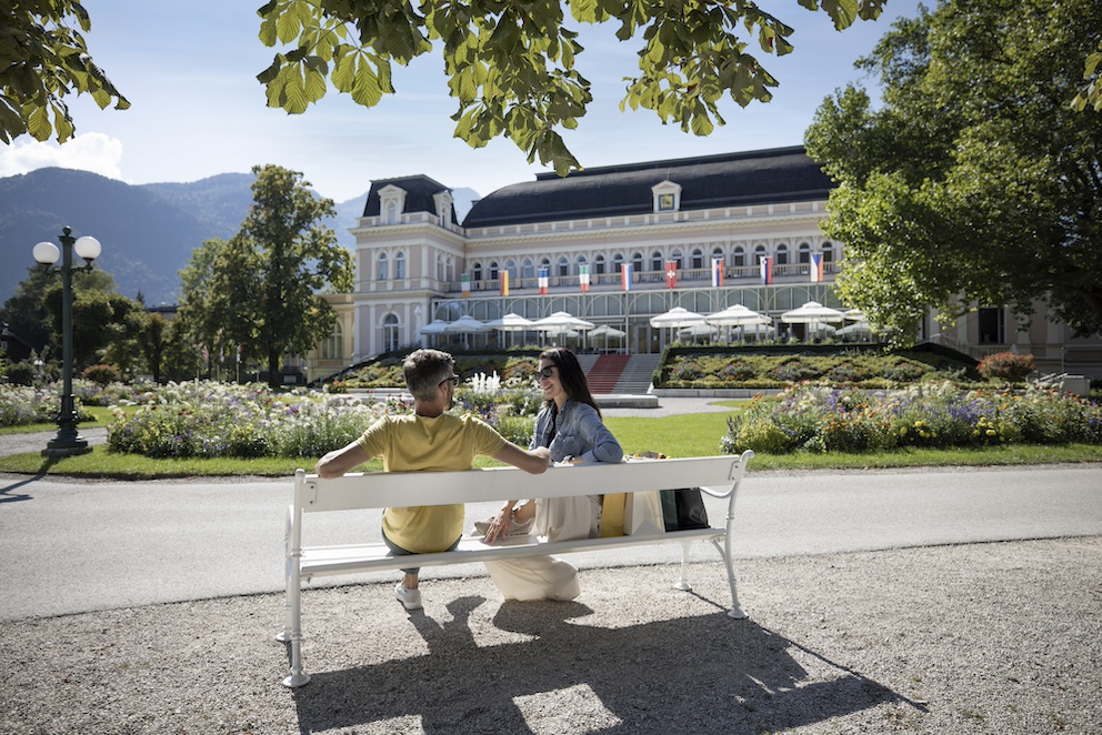 Mann und Frau auf einer Parkbank im Kurpark in Bad Ischl, hinter ihnen das historische Kongress- und Theaterhaus.