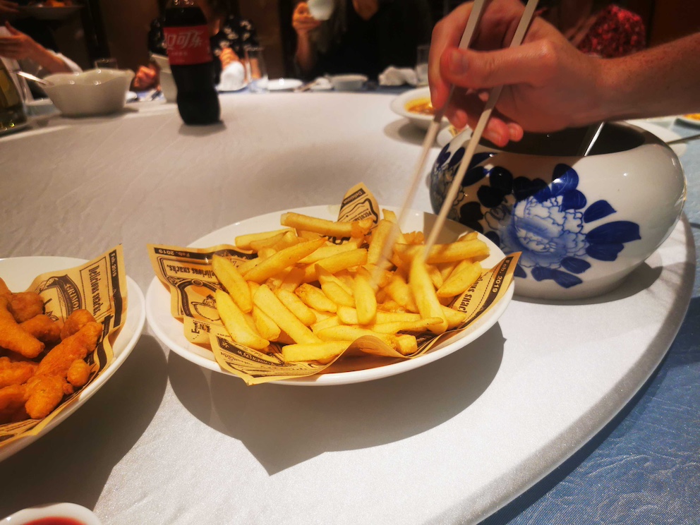 Eine Hand isst Pommes mit Stäbchen an einem runden Esstisch, der westliche und chinesische Elemente vereint.