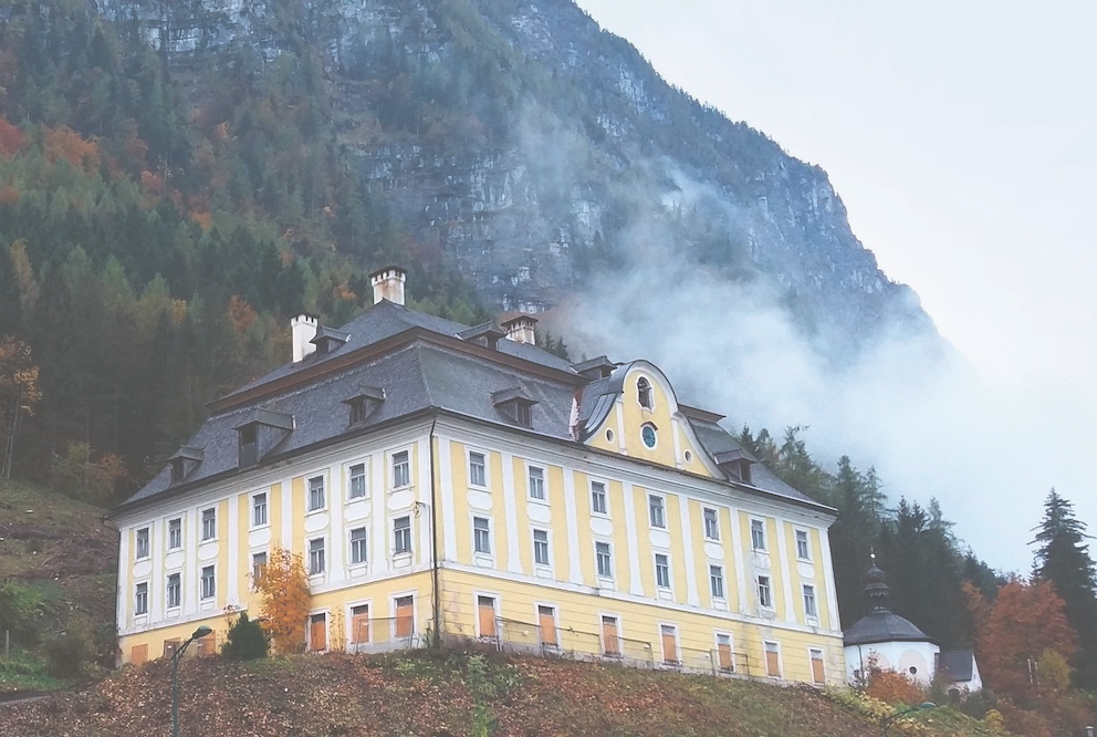 Gelbes historisches Gebäude im Salzkammergut, das vor nebelverhangenen, bewaldeten Bergen steht. Das herbstliche Laub und die dichte Bewaldung rahmen die Szenerie ein.