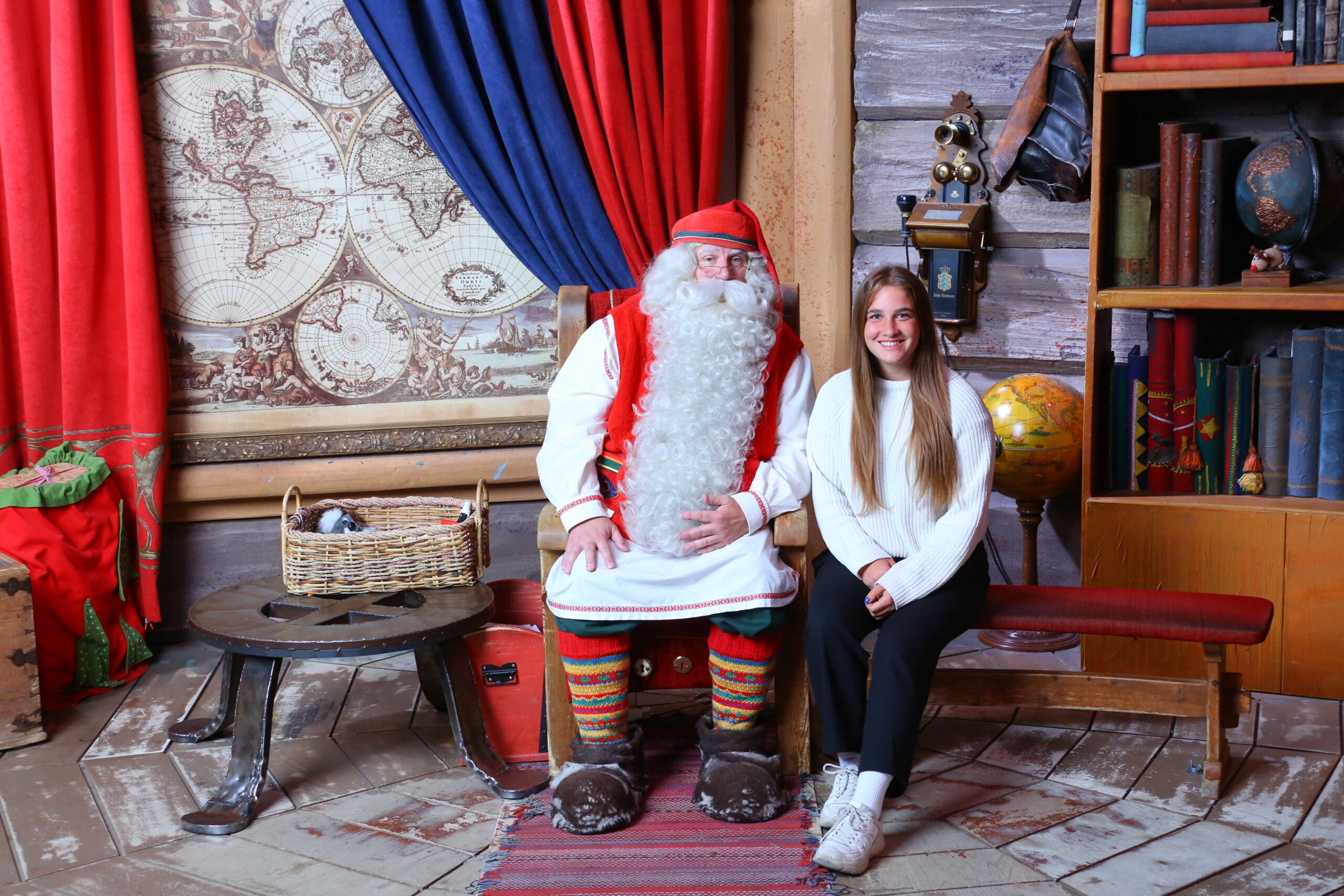Eine Frau Alina Lingg sitzt neben dem Weihnachtsmann im Santa Claus Village in Finnland, umgeben von weihnachtlicher Dekoration.