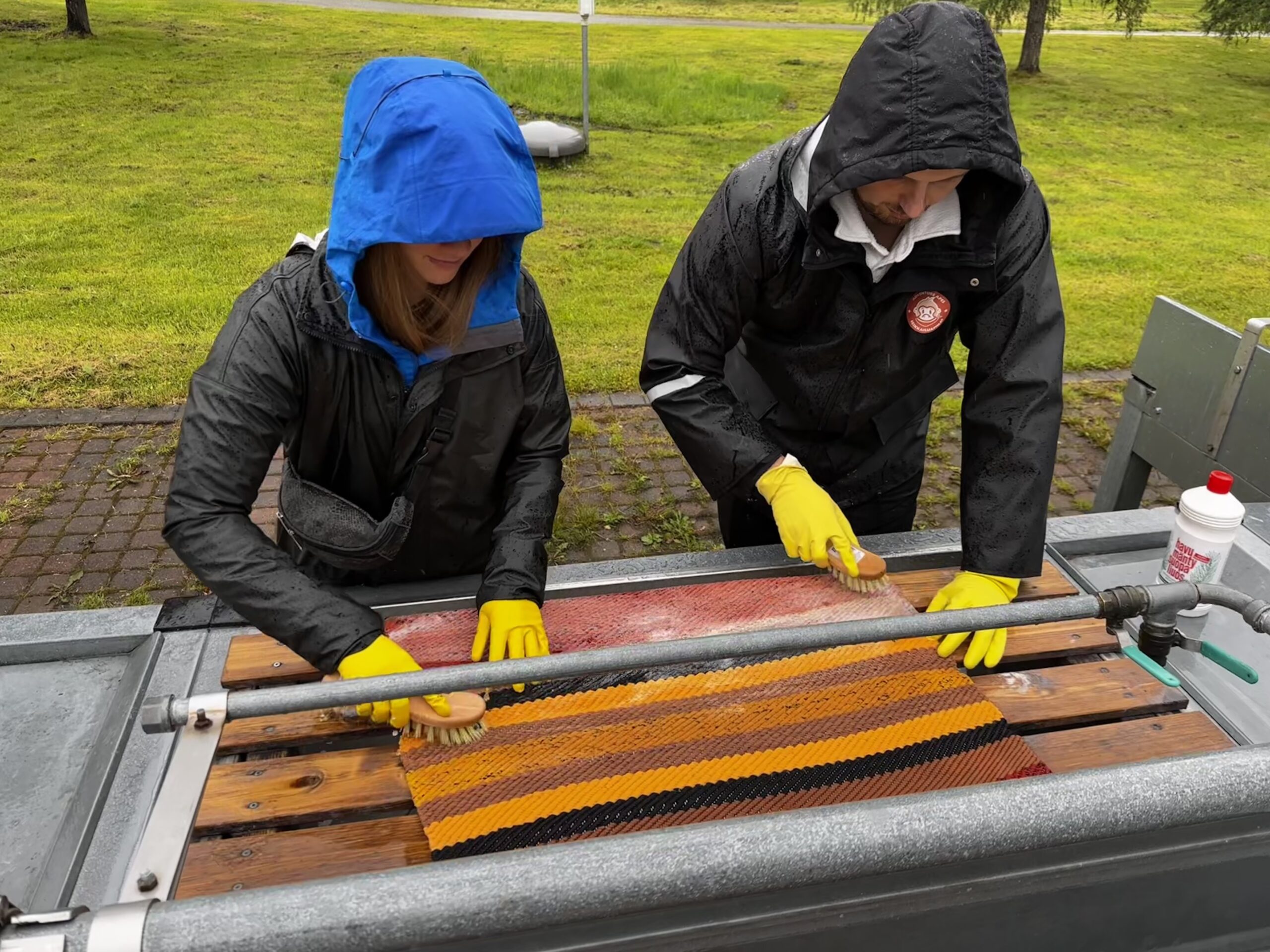 Zwei Personen waschen einen Teppich im Freien in Finnland, gekleidet in Regenjacken und gelben Handschuhen.