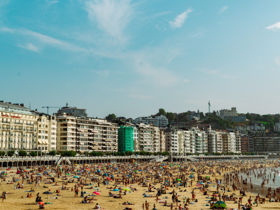 Ein belebter Strand in San Sebastián, Spanien, mit zahlreichen Badegästen. Im Hintergrund sind hohe Wohngebäude und grüne Hügel zu sehen, die sich unter einem klaren blauen Himmel erstrecken.