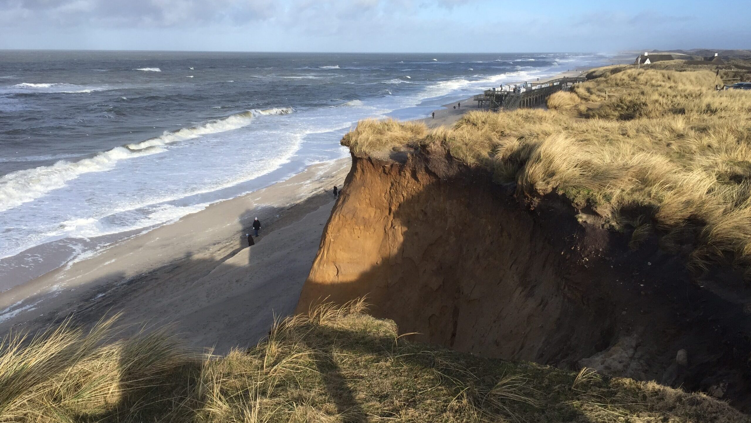 Steilküste auf Sylt mit Blick auf die Nordsee, sandige Dünen und Gräser, herbstlicher Himmel und hohe Wellen bei windigem Wetter.