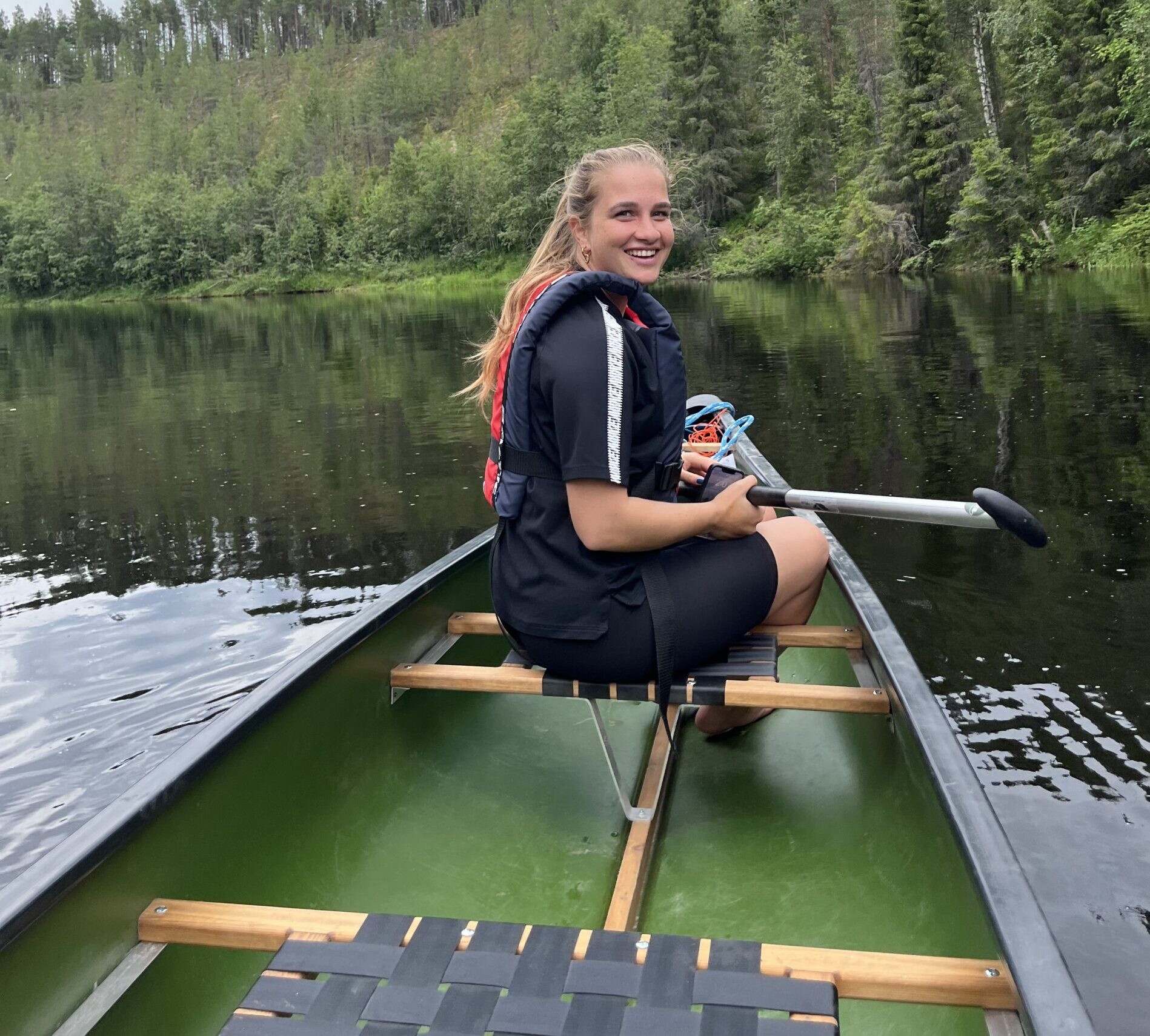 ALT-Text: "Frau sitzt in einem Kanu auf einem Fluss, umgeben von dichtem Wald, unter einem wolkigen Himmel in Finnland.