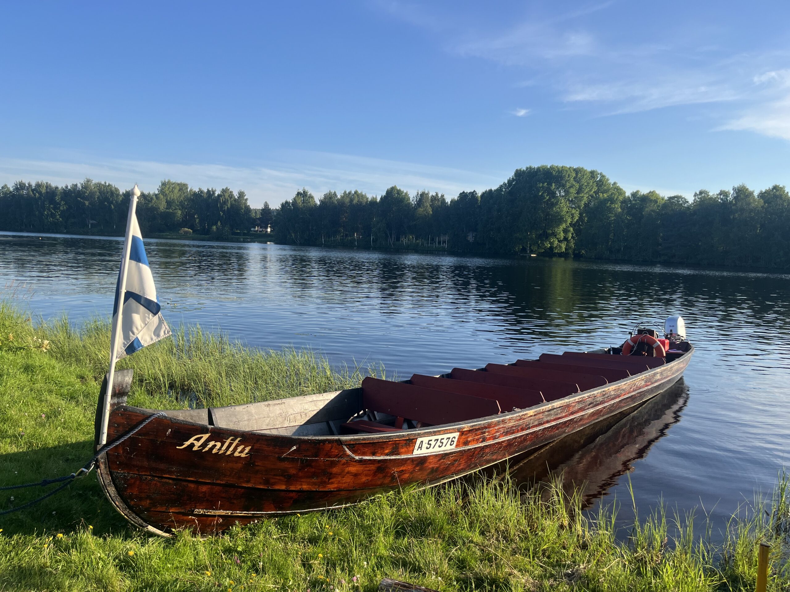 Holzboot mit finnischer Flagge am Ufer eines Sees in Finnland bei sonnigem Wetter.