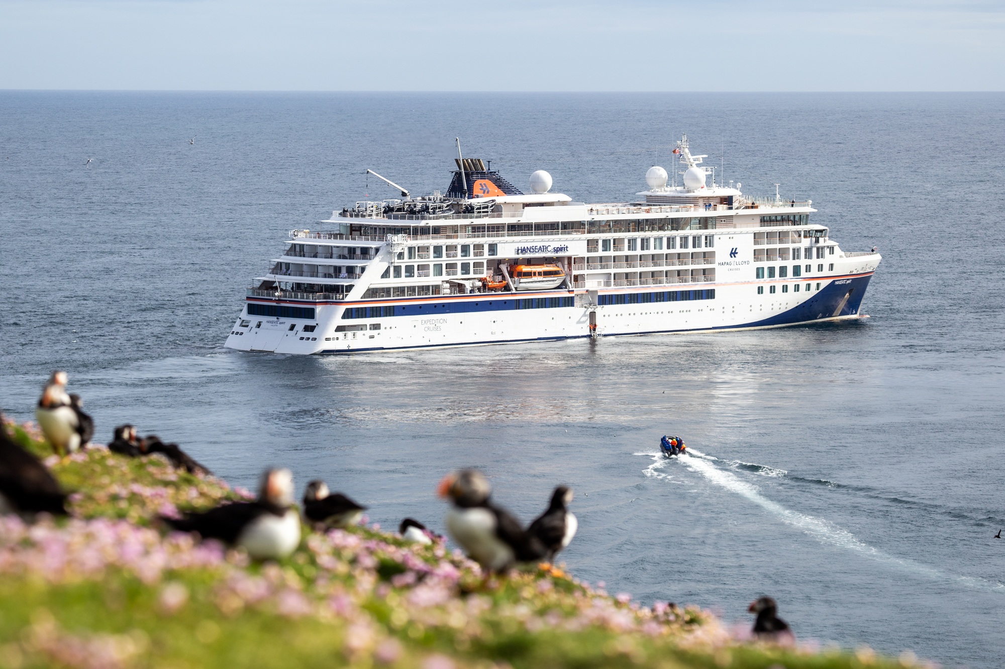 Ein Expeditionsschiff und ein Zodiac auf dem Meer, im Vordergrund Papageitaucher auf einem Felsen.