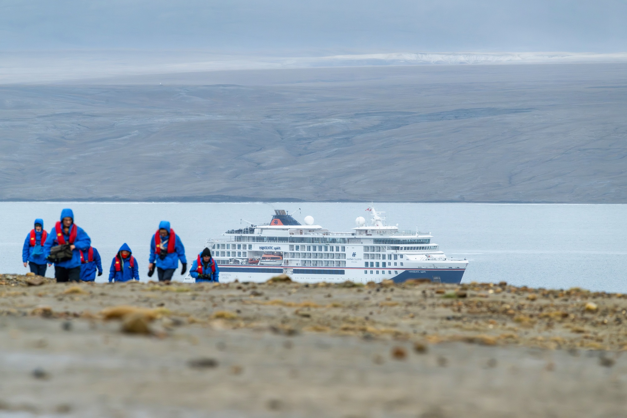 Reisende in blauen Parkas an einer arktischen Küste, dahinter ein Expeditionsschiff.