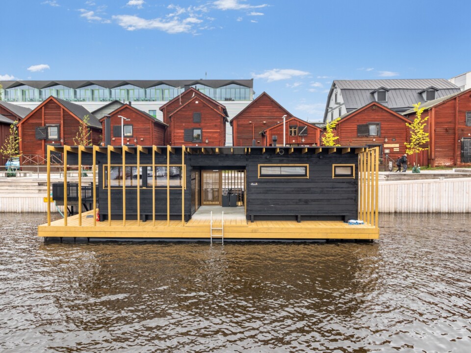 Die schwarze Hehku-Sauna auf einer schwimmenden Plattform vor roten Holzhäusern am Wasser in Oulu.