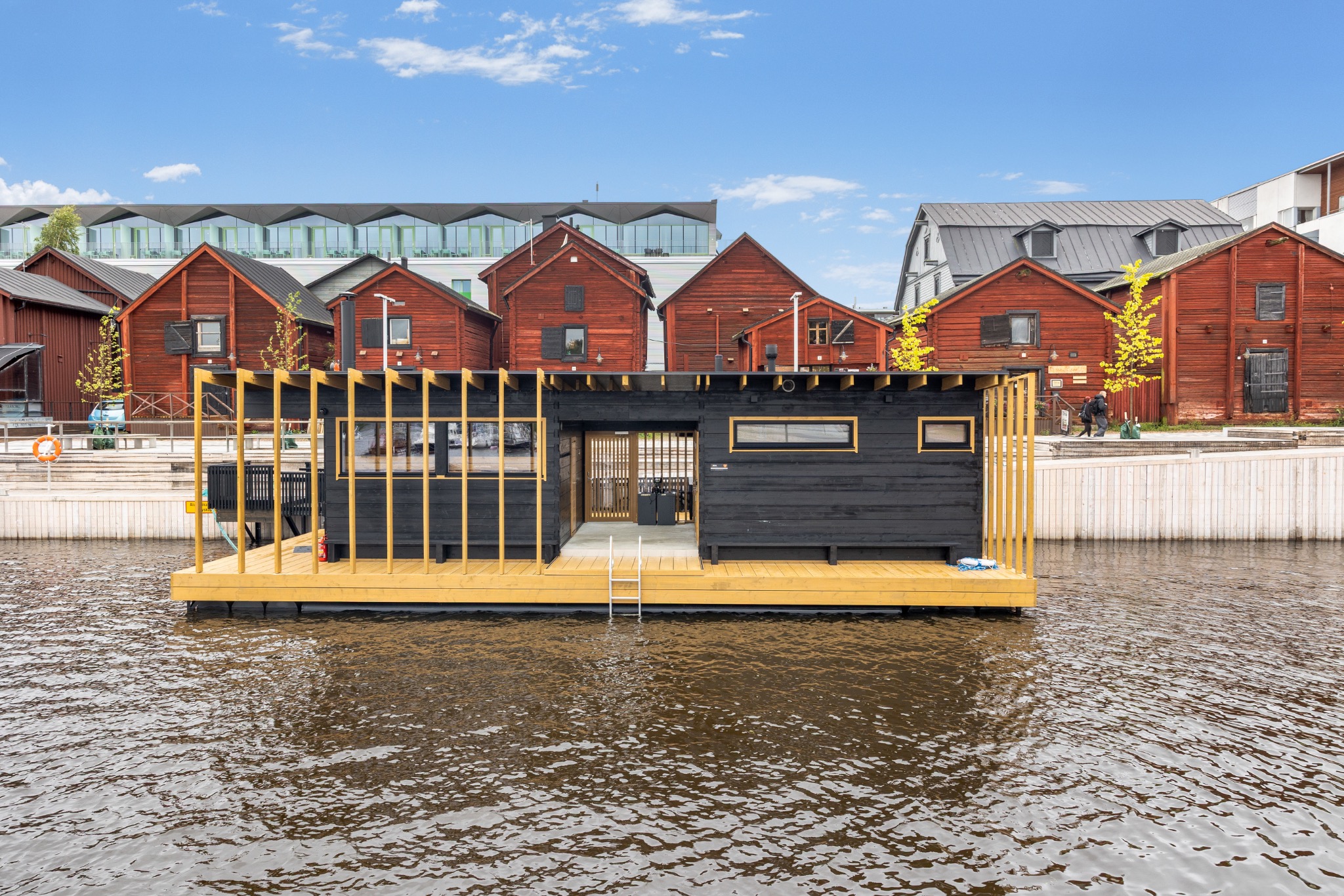 Die schwarze Hehku-Sauna auf einer schwimmenden Plattform vor roten Holzhäusern am Wasser in Oulu.