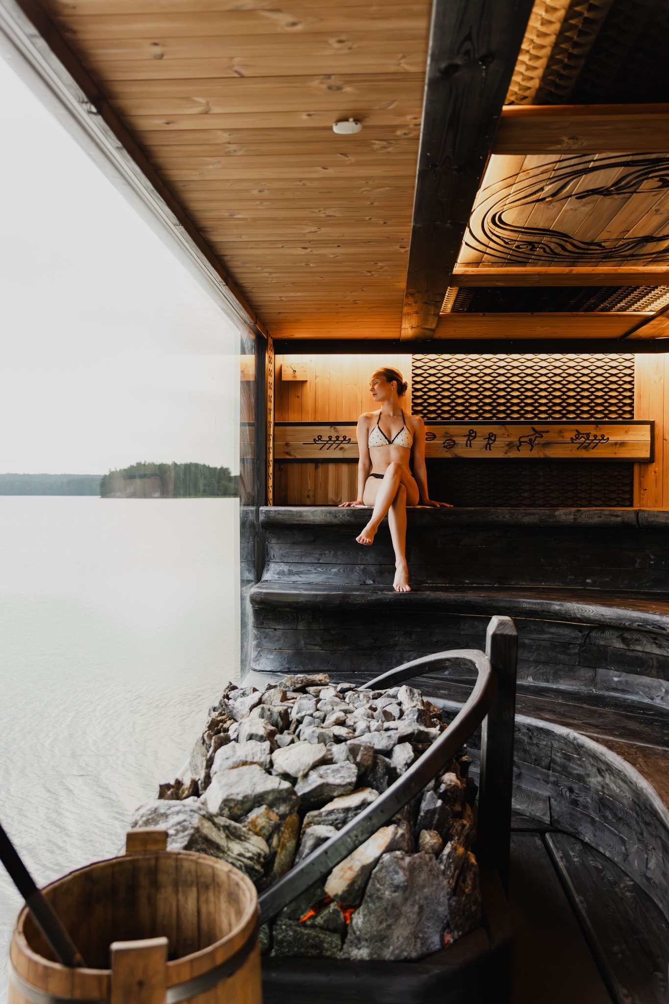 Eine Besucherin sitzt in einer holzverkleideten Sauna vor einem großen Fenster mit Seeblick.