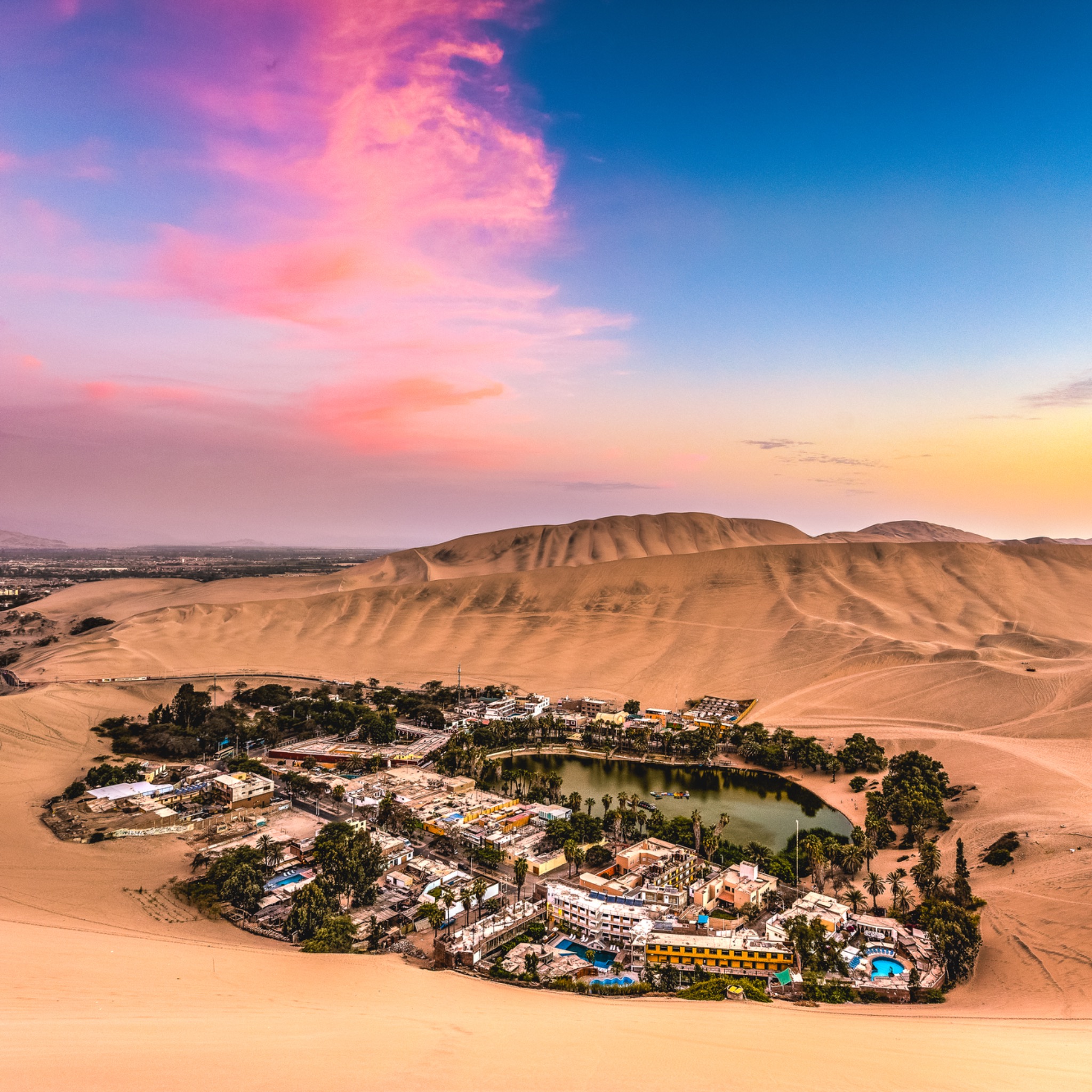 Die Oase Huacachina mit grüner Lagune zwischen hohen Sanddünen bei Sonnenuntergang.