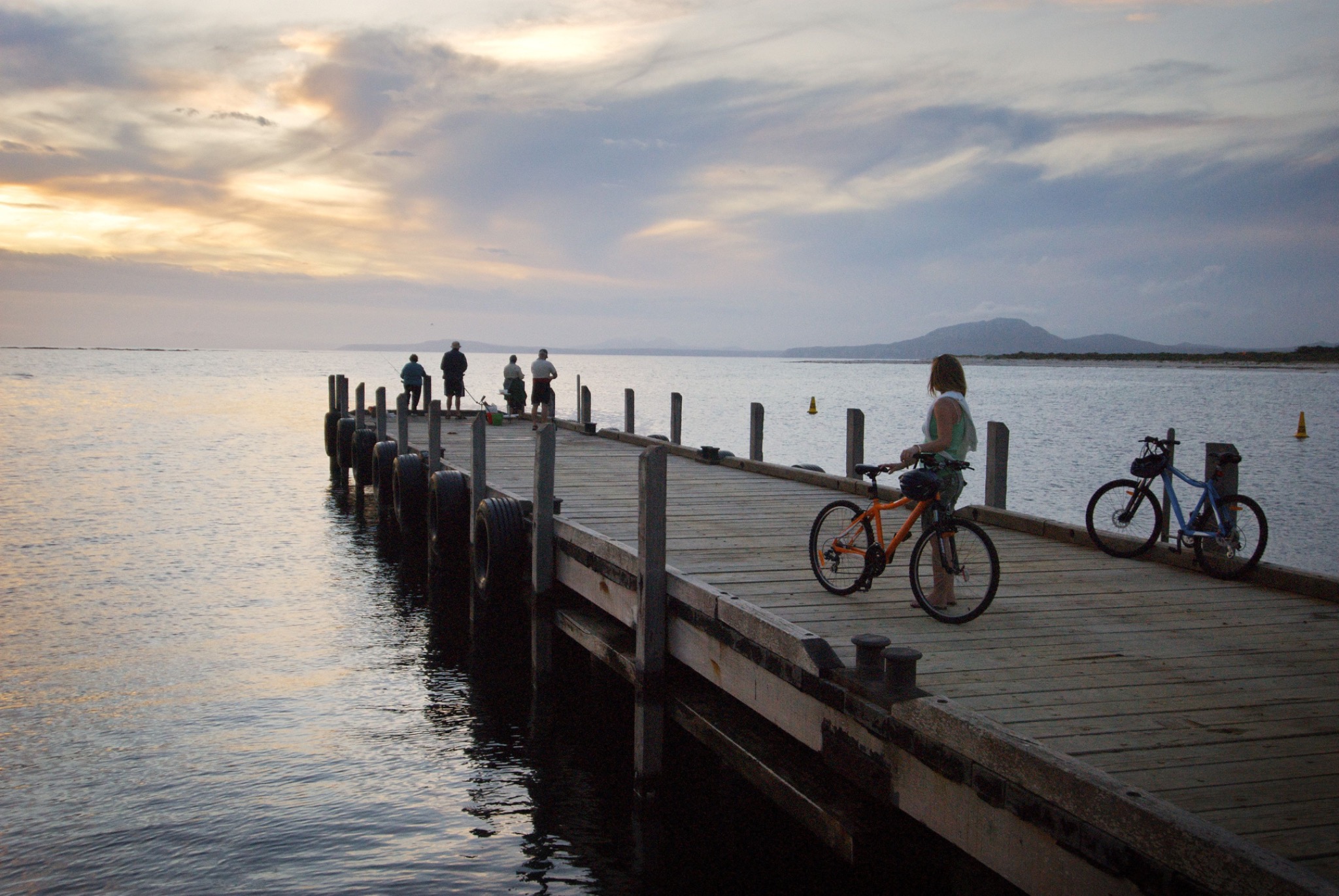 Menschen mit Fahrrädern auf einem Steg am Wasser im Abendlicht.