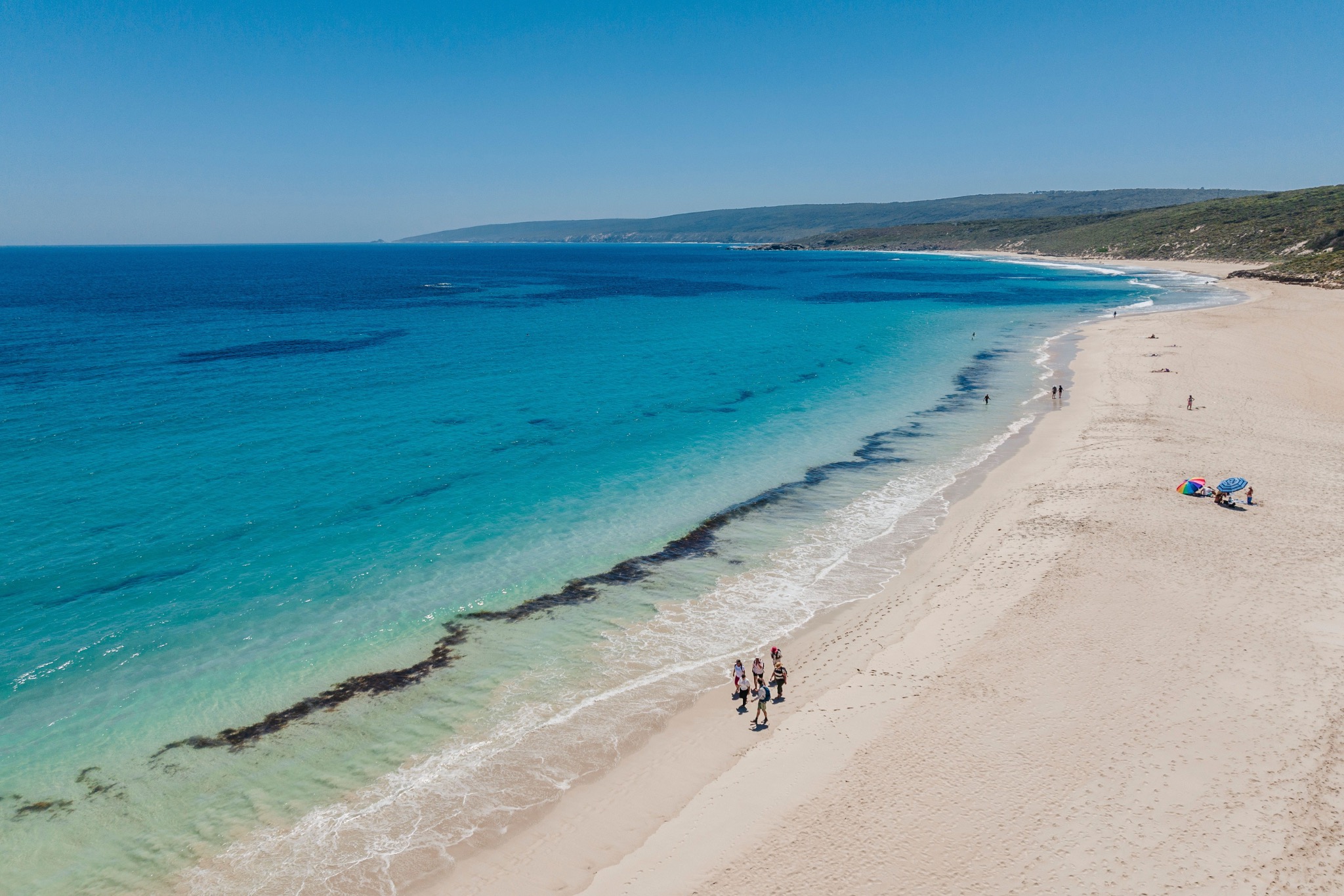 Luftaufnahme eines weißen Sandstrandes mit Spaziergängern an türkisblauem Meer.